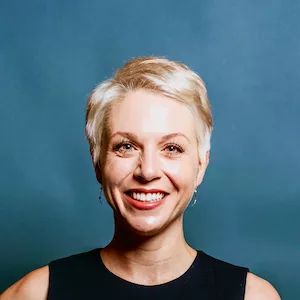 A woman with short blonde hair is wearing a sleeveless black top. She is positioned against a plain blue background, creating a professional and minimalistic visual. The image emphasizes simplicity and elegance.